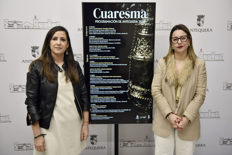 Dos mujeres representantes del ayuntamiento de Antequera posan al lado del cartel de la programación de la Cuaresma de Antequera 2024.