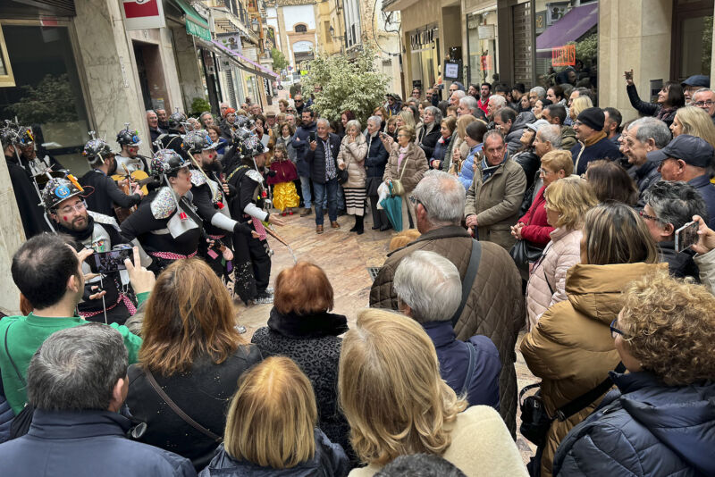 Un gran grupo de personas forman un corrillo para dejar en el centro a otro grupo de chicos y chicas disfrazados. Todos están en una de las calles del lugar rodeado de tiendas. Un ambiente muy divertido.