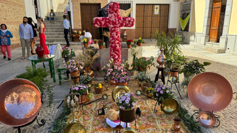 XXXI Concurso de Cruces de mayo 2024. Cruz decorada con flores y vista en las calles del pueblo, escenario con decoraciones de madera, tela y macetas, cacharros de bronce y otros adornos.